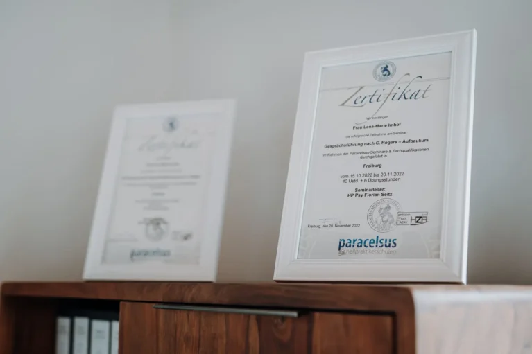 Zwei eingerahmte Zertifikate auf einem Holzregal, die Qualifikation in Psychotherapie zeigen.