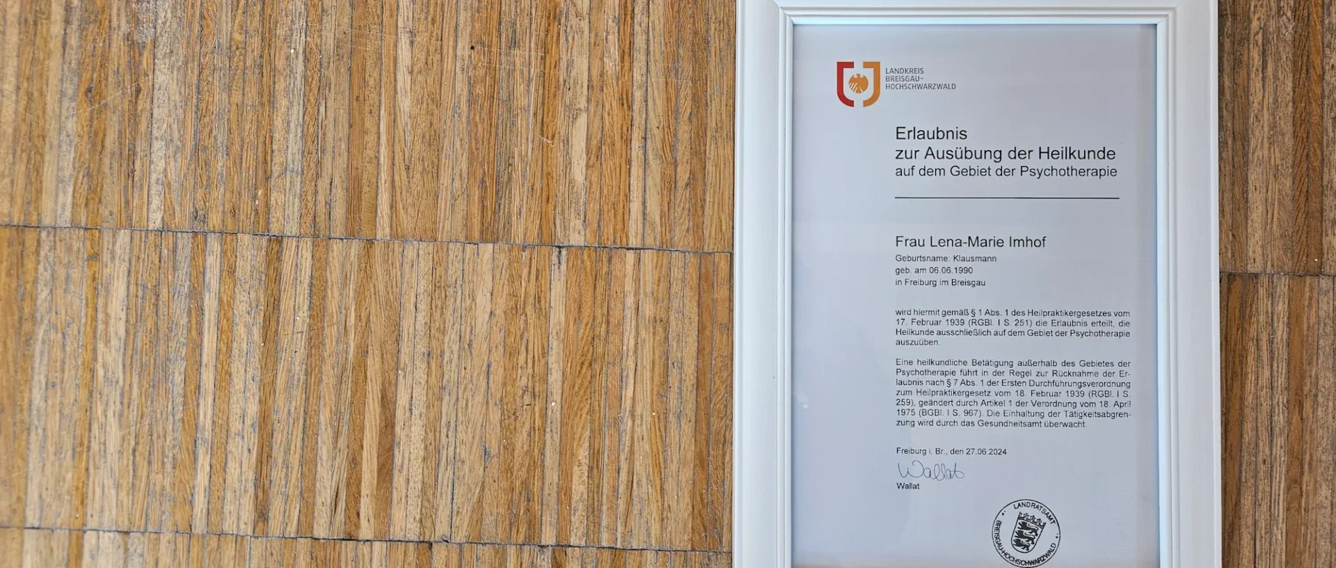 Heilpraktiker-Erlaubnis für Psychotherapie in einem weißen Rahmen an einer Holzwand.
